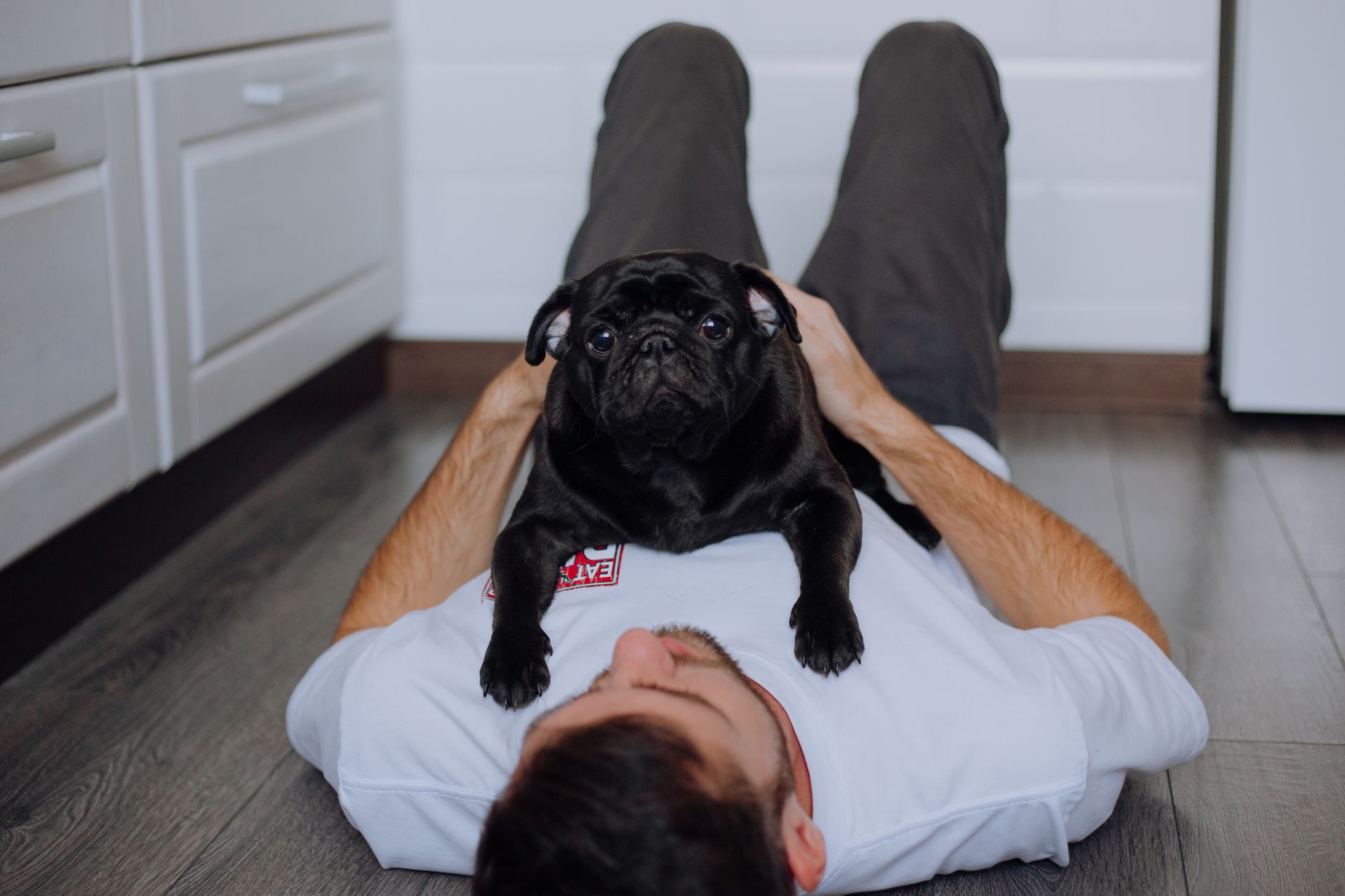 A man lying on the floor with a black pug dog resting on his chest in a kitchen setting.