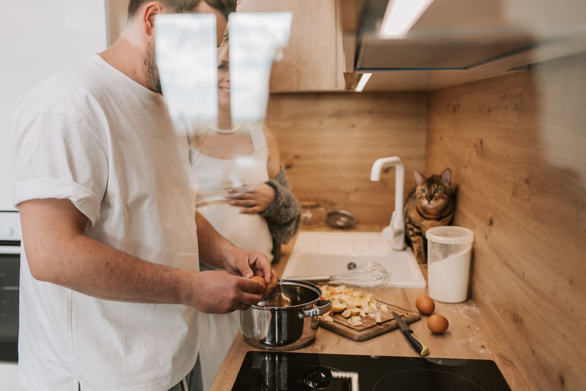A pregnant woman and man cook together in a cozy kitchen, accompanied by their pet cat.